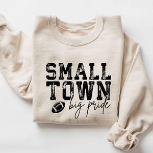 May include: A cream-colored sweatshirt with the words "SMALL TOWN" above "big pride" in black text. A football graphic is between the phrases. The sleeves are rolled up.