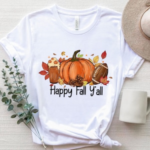以下が含まれることがあります： 秋をテーマにしたデザインの白いTシャツ。オレンジ色のカボチャ、アメフトボール、コーヒー、紅葉のイラストが描かれています。「Happy Fall Y'all」の文字が下にプリントされています。柔らかい素材のシャツです。