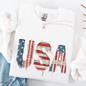 May include: A white sweatshirt with the word "USA" appliqued on the front. The letters are made from a distressed American flag design with red and white stripes and blue stars. A silver necklace with a small pendant rests on the sweatshirt.