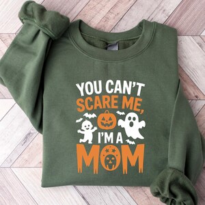 Może przedstawiać: Ciemnozielona bluza z napisem "YOU CAN'T SCARE ME, I'M A MOM" w kolorze białym i pomarańczowym. Projekt zawiera grafiki związane z Halloween: ducha, dynię i nietoperze.