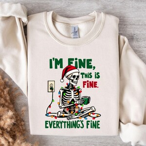 Może przedstawiać: Kremowa bluza z zabawną grafiką. Projekt przedstawia szkielet w czapce Świętego Mikołaja, otoczony lampkami choinkowymi i trzymający kubek. Tekst brzmi "I'm Fine, This is Fine, Everything's Fine."