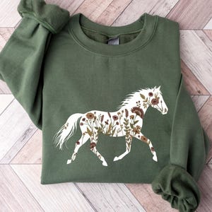 Może przedstawiać: Zielona bluza w kolorze oliwkowym z białym kształtem konia wypełnionym kwiatowymi wzorami w odcieniach brązu i beżu. Koń jest przedstawiony w biegu, tworząc wzór inspirowany naturą. Bluza ma długie rękawy.