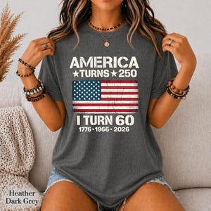 May include: A heather dark grey short-sleeve t-shirt featuring white text that reads AMERICA TURNS 250, followed by a distressed American flag graphic, and the text I TURN 60 with the years 1776, 1966, and 2026 printed in white below the flag.