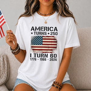 May include: A white short-sleeve t-shirt featuring a graphic of the American flag with the text AMERICA TURNS 250, I TURN 60, and the years 1776 - 1966 - 2026 printed in black. The design has a distressed, weathered appearance on the flag graphic.