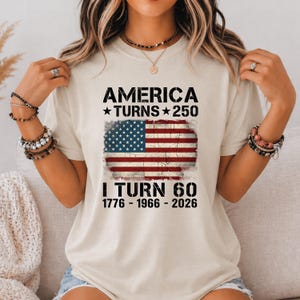 May include: A beige short-sleeve t-shirt featuring a distressed graphic of the American flag. The text reads: AMERICA TURNS 250, I TURN 60, 1776 - 1966 - 2026. The design is centered on the chest in black lettering with a vintage, weathered appearance.