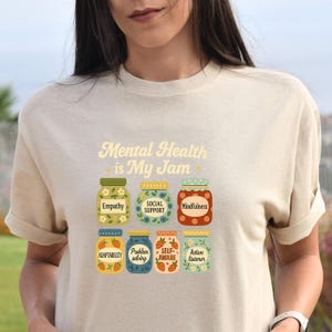 May include: Beige t-shirt with the text "Mental Health is My Jam" and illustrations of jam jars labeled with words like "Empathy," "Social Support," and "Mindfulness." The design features colorful graphics and a playful font.