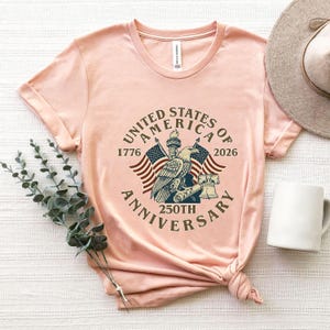 May include: A light pink t-shirt featuring a graphic commemorating the 250th anniversary of the United States. The design includes an eagle, flags, and the text "United States of America 250th Anniversary 1776-2026."