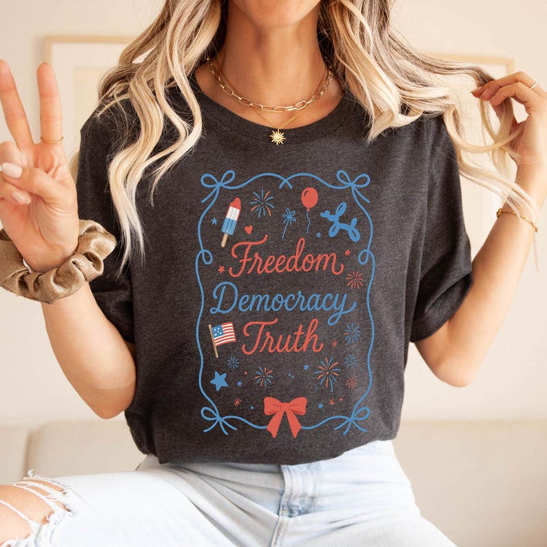 Puede incluir: Camiseta gris oscuro con un dise&ntilde;o patri&oacute;tico. El dise&ntilde;o incluye las palabras "Freedom Democracy Truth" en rojo y azul, junto con ilustraciones de fuegos artificiales, un animal de globo, un helado y una bandera estadounidense.