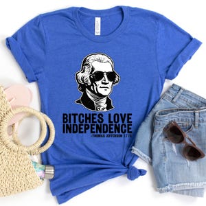 May include: A blue t-shirt with a black and white illustration of Thomas Jefferson wearing sunglasses. The text "Bitches Love Independence -Thomas Jefferson 1776" is printed on the shirt.