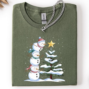 Puede incluir: Camiseta verde oliva con un diseño de muñecos de nieve apilados, un árbol de Navidad y una estrella dorada. Los muñecos de nieve llevan sombreros y bufandas de colores. Una cadena plateada está sobre la camiseta.