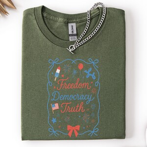 Puede incluir: Camiseta verde oliva con la frase "Freedom Democracy Truth" en rojo y azul. El dise&ntilde;o incluye un helado, fuegos artificiales, un globo y un lazo. Una cadena de plata descansa sobre la camiseta.