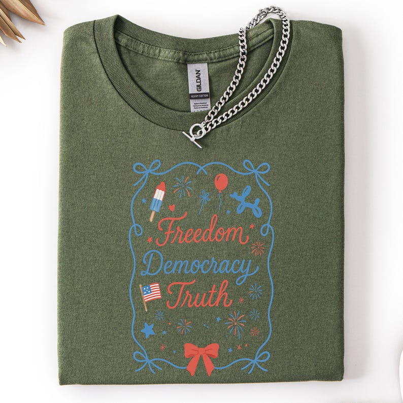 Puede incluir: Camiseta verde oliva con la frase "Freedom Democracy Truth" en rojo y azul. El dise&ntilde;o incluye un helado, fuegos artificiales, un globo y un lazo. Una cadena de plata descansa sobre la camiseta.
