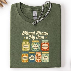May include: Olive green t-shirt with the text "Mental Health is My Jam" and illustrations of jam jars labeled with positive mental health concepts. A silver chain necklace rests on the shirt.