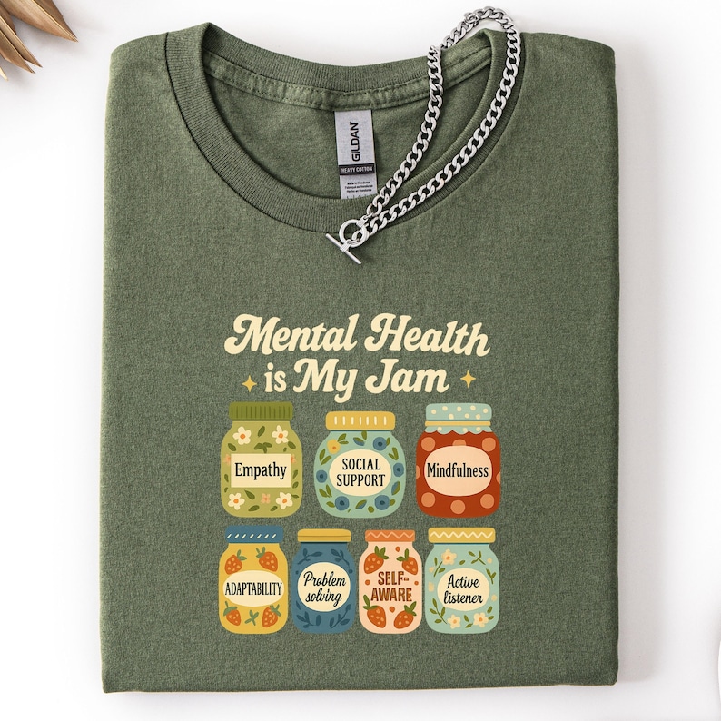May include: Olive green t-shirt with the text "Mental Health is My Jam" and illustrations of jam jars labeled with positive mental health concepts. A silver chain necklace rests on the shirt.