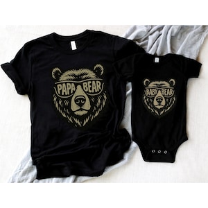 以下が含まれることがあります： クマのグラフィックが描かれた黒いTシャツ2枚。1枚は「PAPA BEAR」、もう1枚は「BABY BEAR」と書かれています。どちらのクマのイラストもサングラスをかけています。これらのシャツは、おそらく親と子供用です。