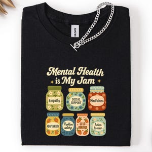 May include: Black t-shirt with the text "Mental Health is My Jam" and illustrations of jam jars labeled with positive mental health concepts like "Empathy" and "Mindfulness". A silver chain is draped over the shirt.
