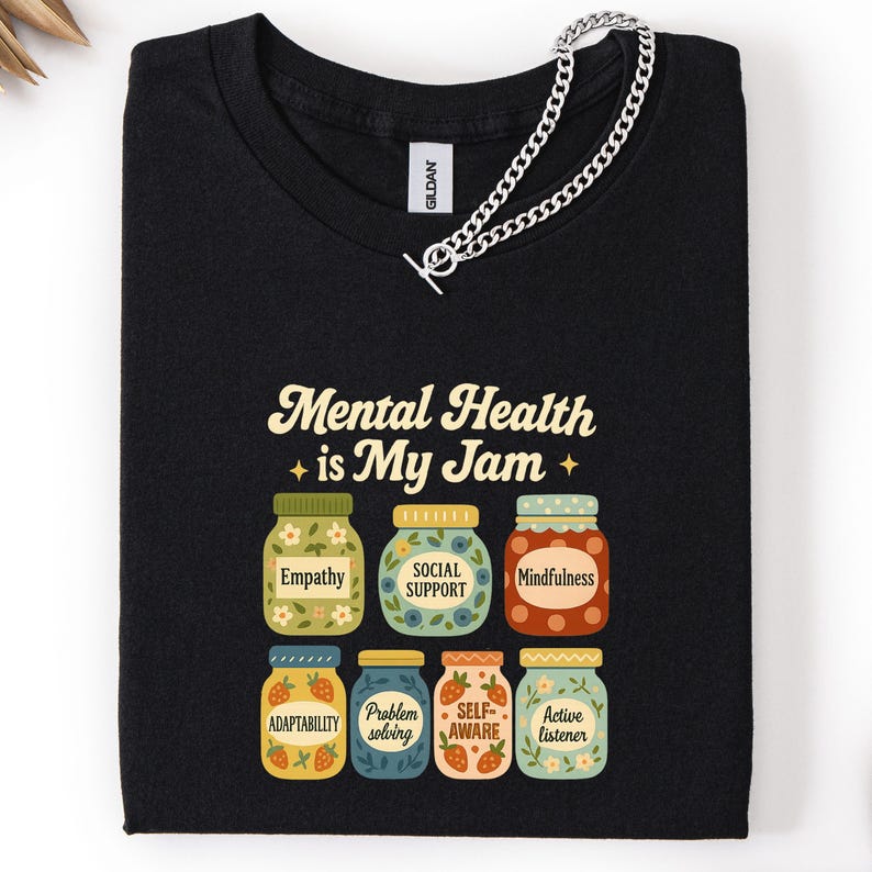 May include: Black t-shirt with the text "Mental Health is My Jam" and illustrations of jam jars labeled with positive mental health concepts like "Empathy" and "Mindfulness". A silver chain is draped over the shirt.