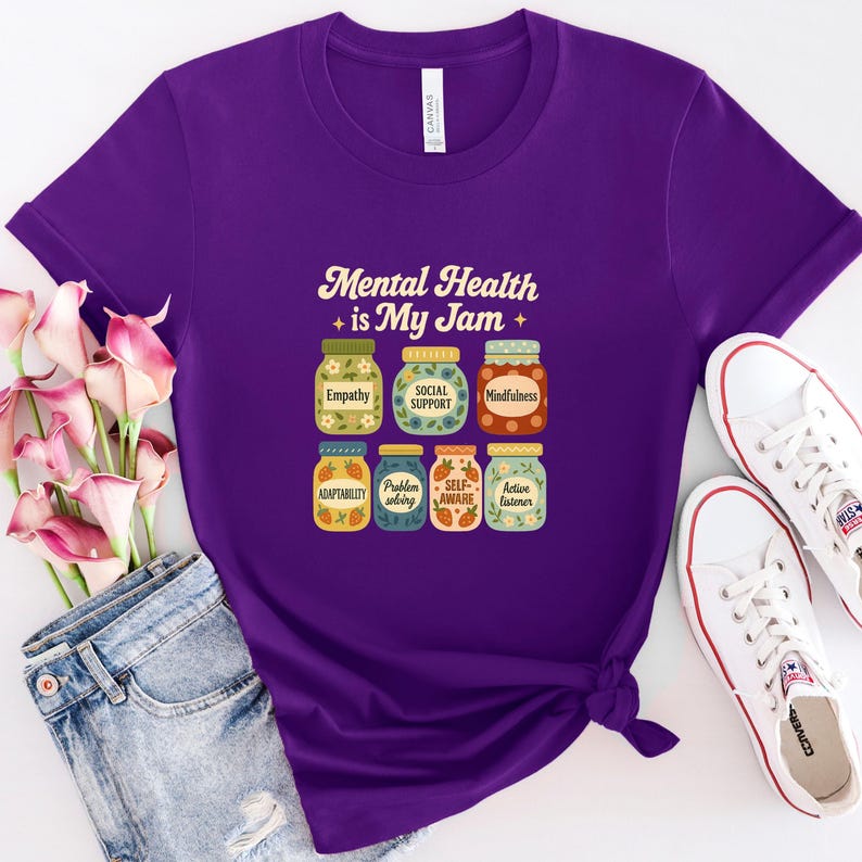 May include: A purple t-shirt with the text "Mental Health is My Jam" and illustrations of jam jars labeled with positive mental health concepts like "Empathy" and "Mindfulness". White sneakers and blue jeans are also in the image.