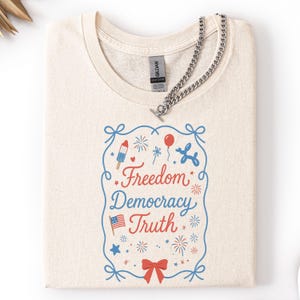 Puede incluir: Camiseta color crema con un dise&ntilde;o gr&aacute;fico que dice "Freedom Democracy Truth" en rojo y azul. El dise&ntilde;o incluye fuegos artificiales, un helado, globos y una bandera americana. Un collar de cadena plateada est&aacute; sobre la camiseta.