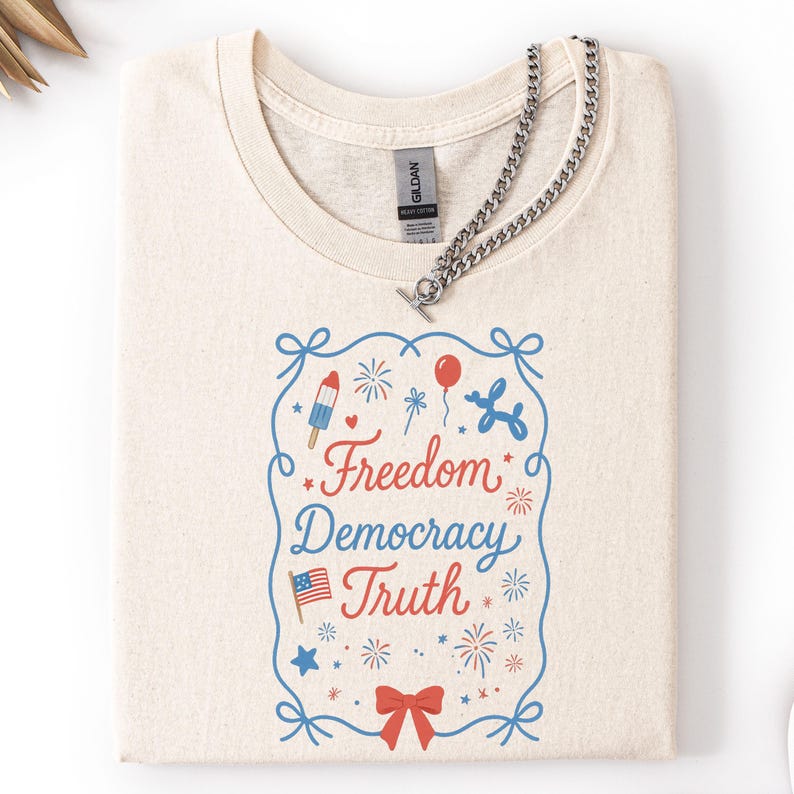 Puede incluir: Camiseta color crema con un dise&ntilde;o gr&aacute;fico que dice "Freedom Democracy Truth" en rojo y azul. El dise&ntilde;o incluye fuegos artificiales, un helado, globos y una bandera americana. Un collar de cadena plateada est&aacute; sobre la camiseta.