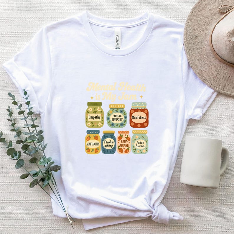 May include: White t-shirt featuring the text "Mental Health is My Jam" above illustrations of jars labeled with positive mental health concepts like "Empathy" and "Mindfulness." A neutral-colored hat and a white mug are also visible.