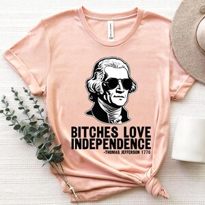 May include: A peach-colored t-shirt with a black and white illustration of a man wearing sunglasses. The text on the shirt reads "Bitches Love Independence -Thomas Jefferson 1776".