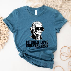 May include: A teal blue t-shirt with a black and white illustration of Thomas Jefferson wearing sunglasses. The text on the shirt reads "Bitches Love Independence -Thomas Jefferson 1776".