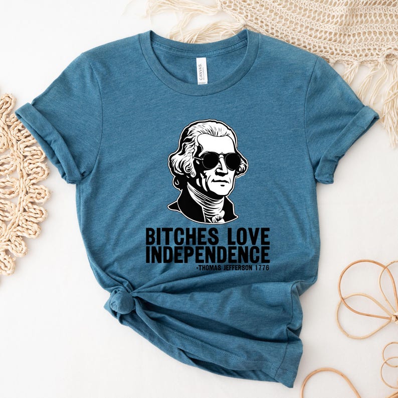 May include: A teal blue t-shirt with a black and white illustration of Thomas Jefferson wearing sunglasses. The text on the shirt reads "Bitches Love Independence -Thomas Jefferson 1776".