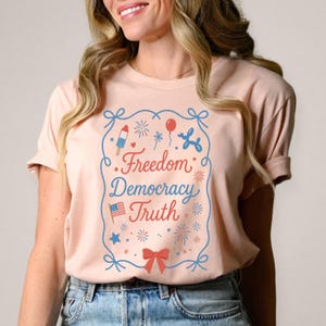 Puede incluir: Camiseta rosa claro con un dise&ntilde;o azul y rojo. El dise&ntilde;o incluye las palabras "Freedom Democracy Truth" e ilustraciones de fuegos artificiales, un perro globo, un helado y una bandera americana.