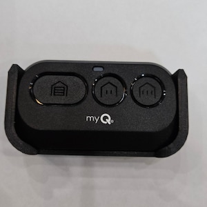 May include: Black myQ garage door opener remote with three buttons and the myQ logo. The buttons include a garage door icon and two house icons. The remote is rectangular with rounded edges.