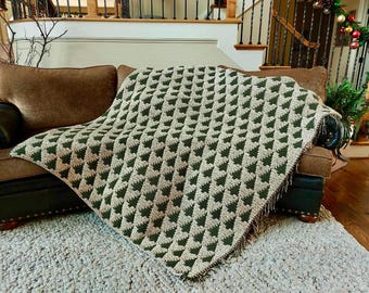 May include: A green and beige knitted throw blanket with a repeating Christmas tree pattern. The blanket is draped over a brown sofa with a matching cushion. The throw has a fringed edge and is placed on a light-coloured rug.