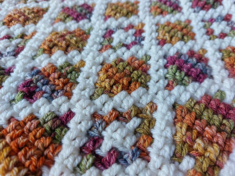 Puede incluir: Primer plano de una manta de ganchillo con un patr&oacute;n floral repetitivo. Las flores son en tonos naranja, rosa, verde y morado, sobre un fondo blanco. La textura es suave y acogedora, ideal para la decoraci&oacute;n del hogar.