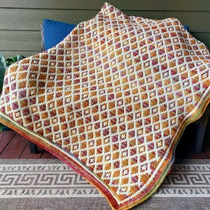 May include: A handmade crochet blanket with a geometric diamond pattern in shades of orange, white, and burgundy. The blanket is draped over a chair and has a colourful border. The blanket measures approximately 152 cm by 152 cm.