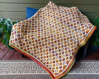 Puede incluir: Una manta de ganchillo hecha a mano con un patrón geométrico de diamantes en tonos naranja, blanco y burdeos. La manta está sobre una silla y tiene un borde colorido. La manta mide aproximadamente 152 cm por 152 cm.