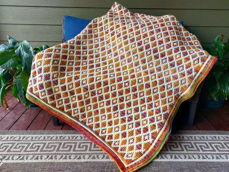 Puede incluir: Una manta de ganchillo hecha a mano con un patr&oacute;n geom&eacute;trico de diamantes en tonos naranja, blanco y burdeos. La manta est&aacute; sobre una silla y tiene un borde colorido. La manta mide aproximadamente 152 cm por 152 cm.