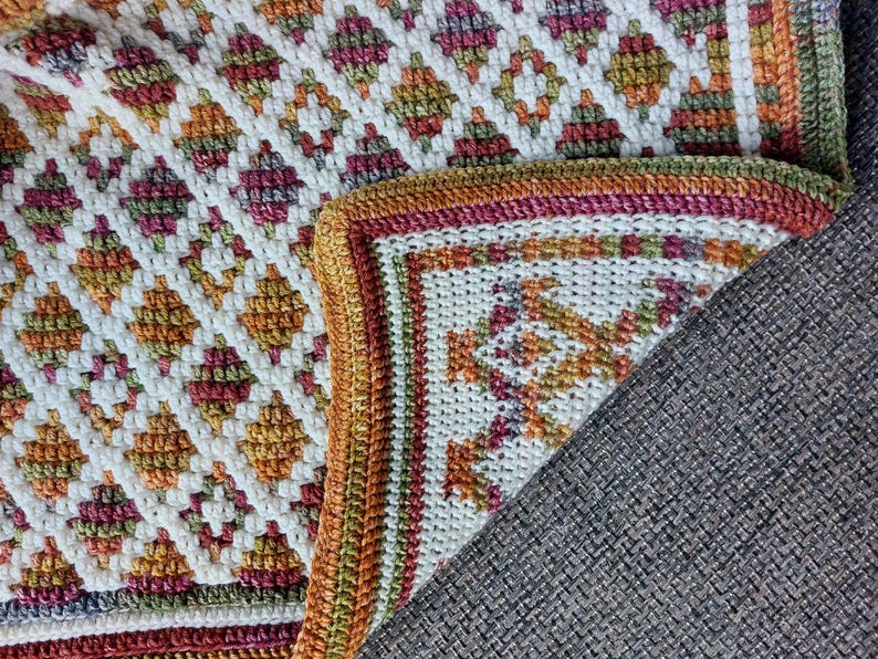 Puede incluir: Una manta de ganchillo hecha a mano con un patr&oacute;n de diamantes geom&eacute;tricos en blanco, naranja, verde y burdeos. La manta tiene un borde detallado con colores y patrones similares. La manta est&aacute; doblada sobre una superficie gris.