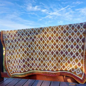 Puede incluir: Manta de ganchillo hecha a mano con un dise&ntilde;o de diamantes en crema, amarillo, naranja y burdeos. El borde de la manta tiene un ribete multicolor. La manta est&aacute; colocada sobre una barandilla de madera, con un cielo azul de fondo.