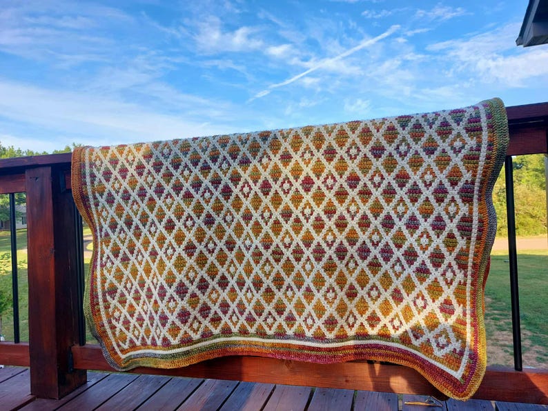 Puede incluir: Manta de ganchillo hecha a mano con un dise&ntilde;o de diamantes en crema, amarillo, naranja y burdeos. El borde de la manta tiene un ribete multicolor. La manta est&aacute; colocada sobre una barandilla de madera, con un cielo azul de fondo.