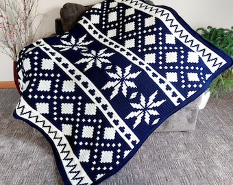 May include: A navy and white knitted blanket with a snowflake and geometric pattern. The blanket features a repeating design of white snowflakes and diamond shapes against a dark blue background. The edges are finished with a white zigzag border.