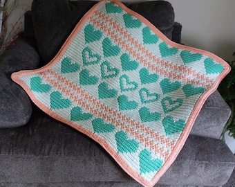 May include: A square, crocheted blanket with a pattern of teal hearts on a white background. The blanket has a diagonal peach-coloured stripe and a peach border. The blanket is folded over a grey armchair.