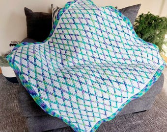 Puede incluir: Una manta de ganchillo con un patrón de diamantes en tonos de blanco, azul, verde y morado. La manta está ribeteada con un borde azul, verde y morado.
