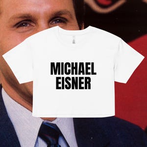 May include: White crop top with the name "MICHAEL EISNER" printed in bold black letters. The shirt has a crew neck and short sleeves. The background features a blurred image of a person in a suit.
