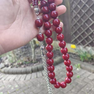 May include: A strand of prayer beads with large, round, deep red beads. The beads are strung together with a silver-colored chain and decorative accents. The top and bottom pieces are a matching deep red color.
