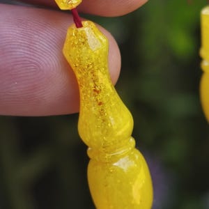 Puede incluir: Primer plano de una cuenta de oración amarilla de un rosario. La cuenta es de color amarillo translúcido con una textura moteada. Un hilo rojo conecta las cuentas. El fondo es verde borroso.