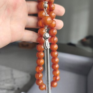 May include: Orange prayer beads with silver-colored accents and a tassel. The beads are round and smooth, strung together with a silver chain. The tassel is made of thin, silver-colored strands.