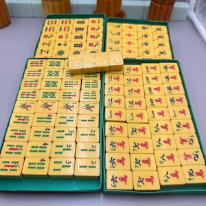 Puede incluir: Un juego de fichas de mahjong amarillas dispuestas en bandejas verdes, con varios símbolos grabados y caracteres chinos en rojo, verde y negro. Una pequeña pila de cuatro fichas amarillas en blanco descansa horizontalmente en el centro.