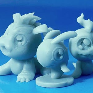 以下が含まれることがあります： 3体の水色のファンタジー生物のフィギュア。フィギュアは大きな目と様式化された特徴を持っています。1つは四足歩行、1つはウサギのような耳、3つ目は頭に炎のような要素があります。青い背景に配置されています。