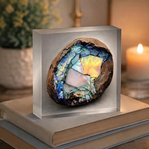 May include: A colorful opal gemstone encased in a clear acrylic block. The opal displays iridescent hues of blue, green, yellow, and orange. The stone is set on a stack of books, with a lit candle and a potted plant in the background.