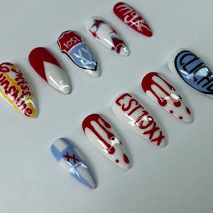 May include: A set of ten long, almond-shaped artificial nails. Each nail is painted with a unique design, including red drips, text, and graphic elements in red, white, blue, and yellow. The designs include the words "lost", "cliche", "miss sunshine", and "est 19xx".