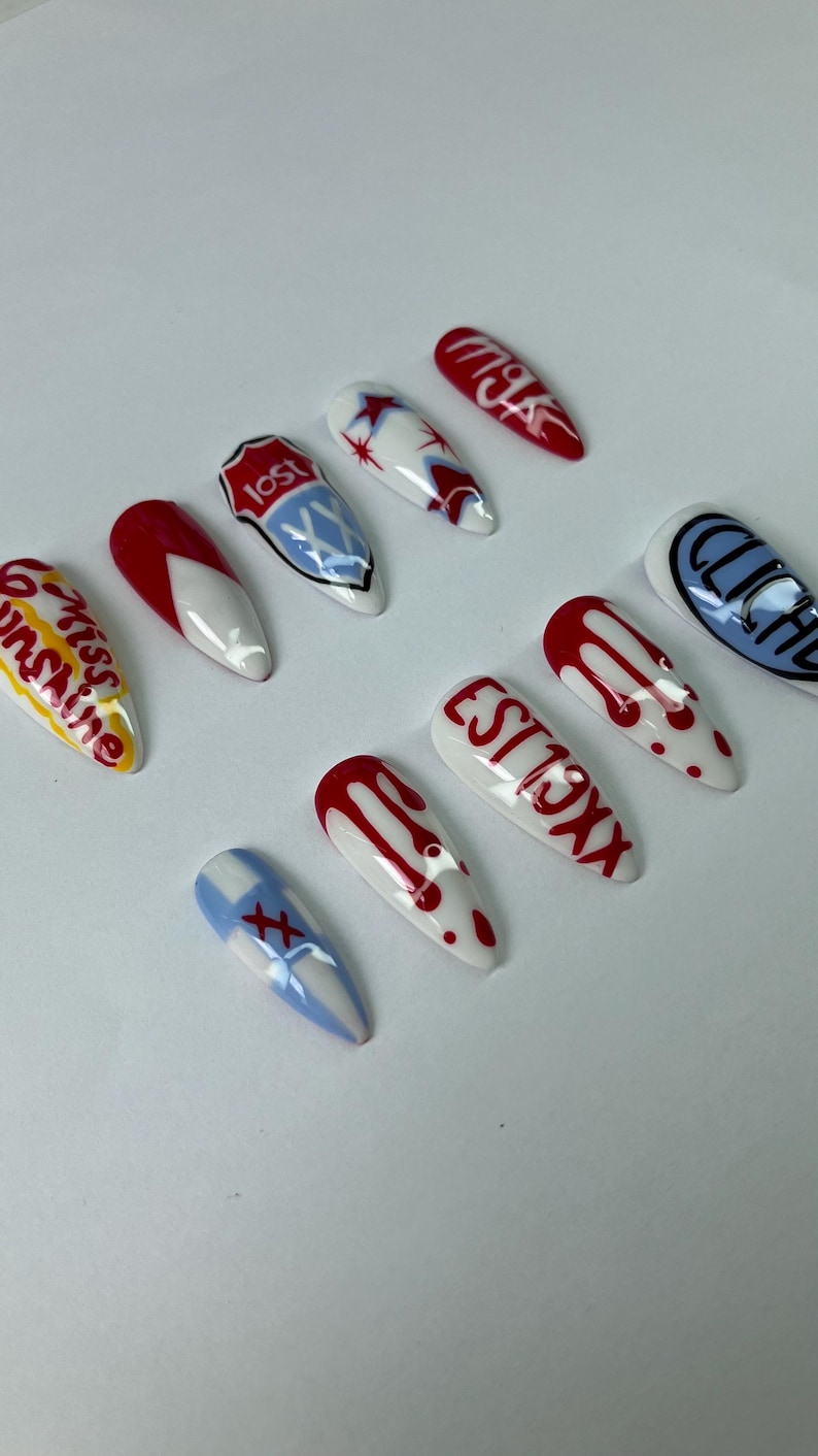 May include: A set of ten long, almond-shaped artificial nails. Each nail is painted with a unique design, including red drips, text, and graphic elements in red, white, blue, and yellow. The designs include the words "lost", "cliche", "miss sunshine", and "est 19xx".